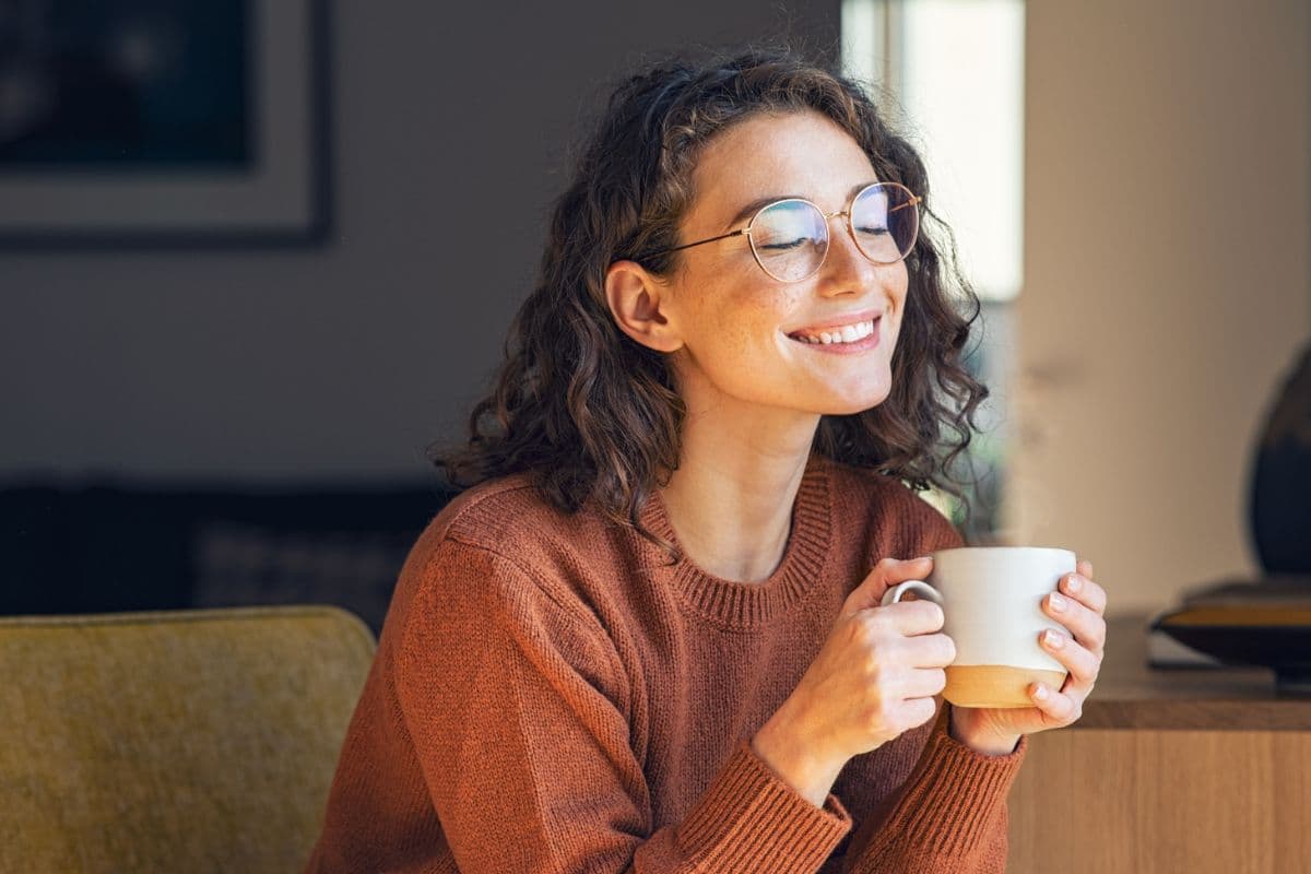 Seorang perempuan sedang tersenyum sembari memegang secangkir minuman hangat