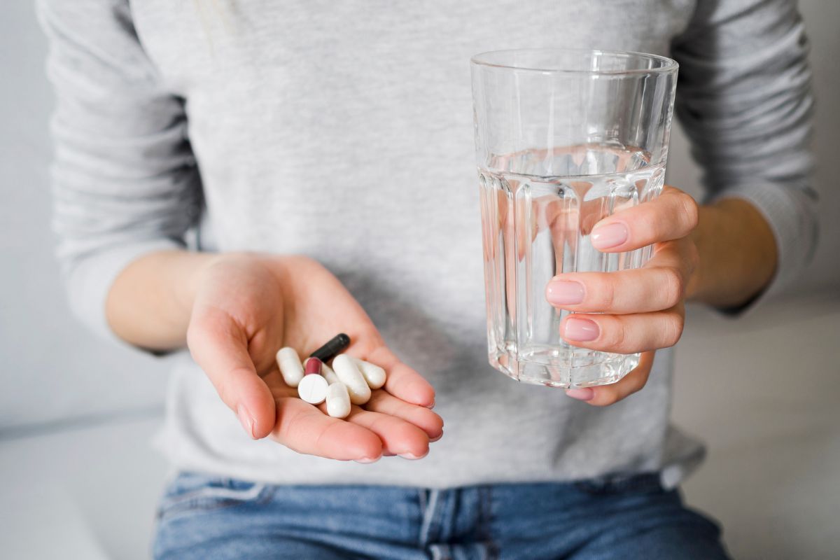 orang yang memegang beberapa obat di satu tangan dan segelas air di tangan yang lain